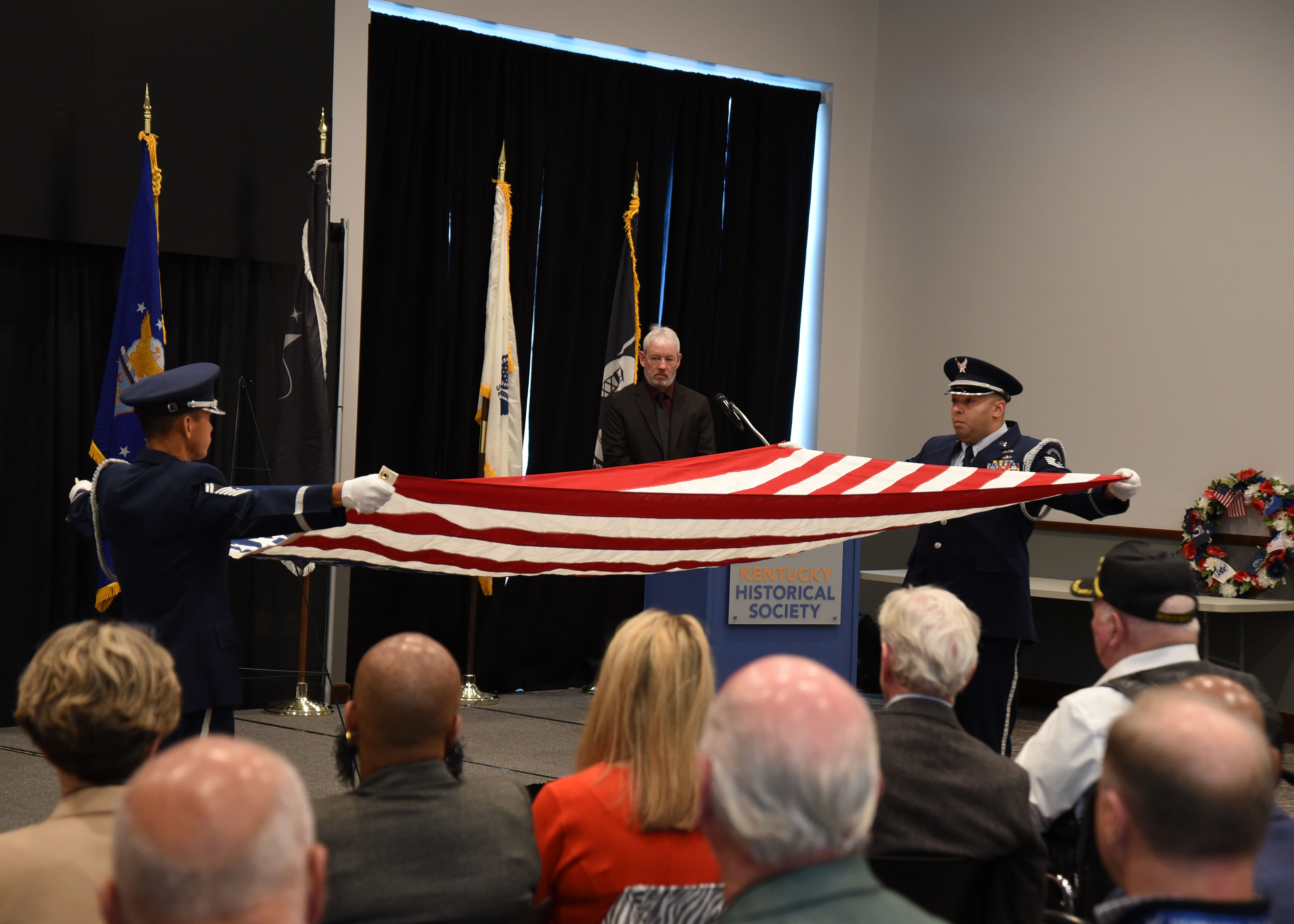 Two uniformed men hold an American flag outstrectched in front of a crowd to display reverence.