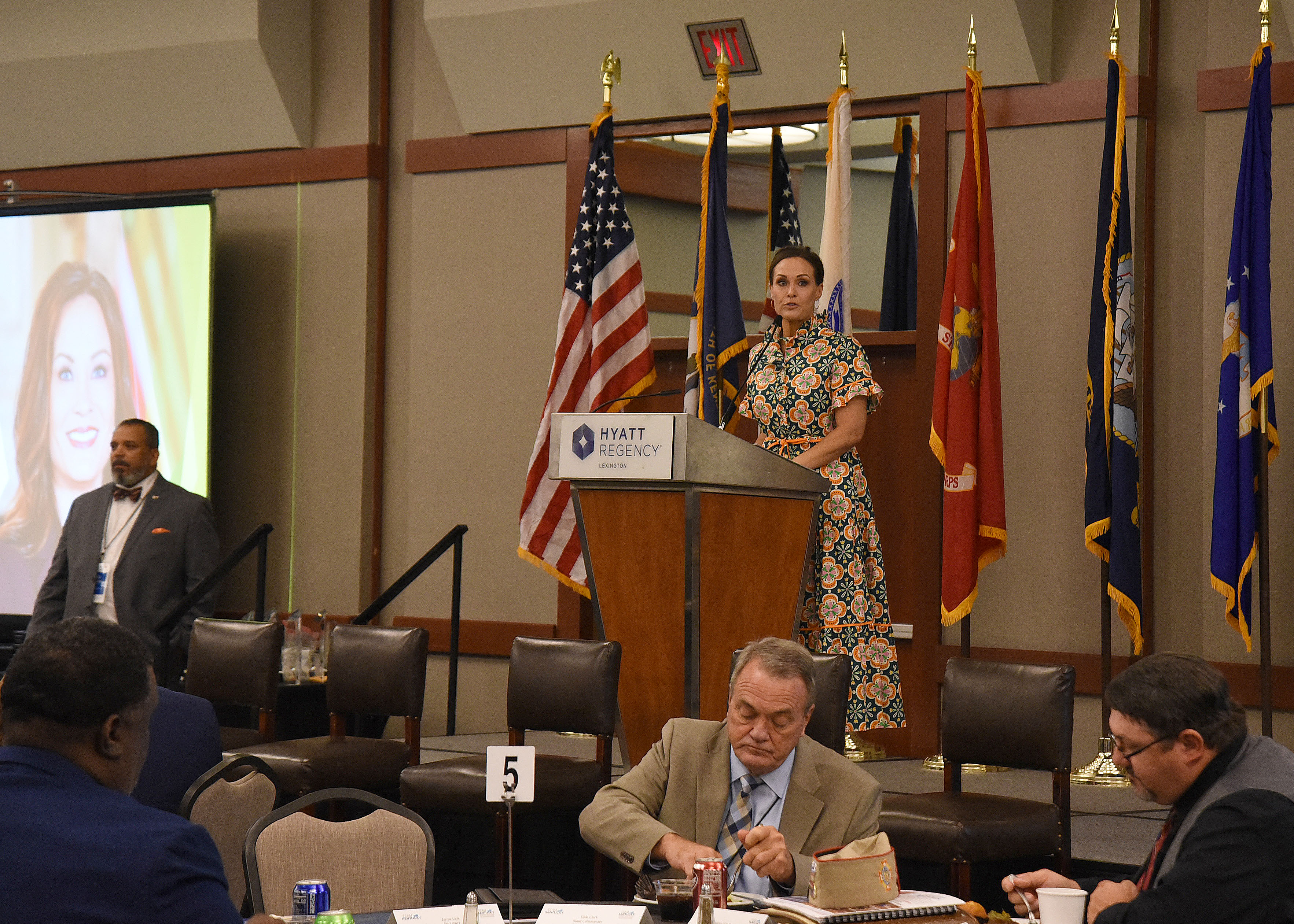 A woman in a dress speaks at a podium in front of a room of people.