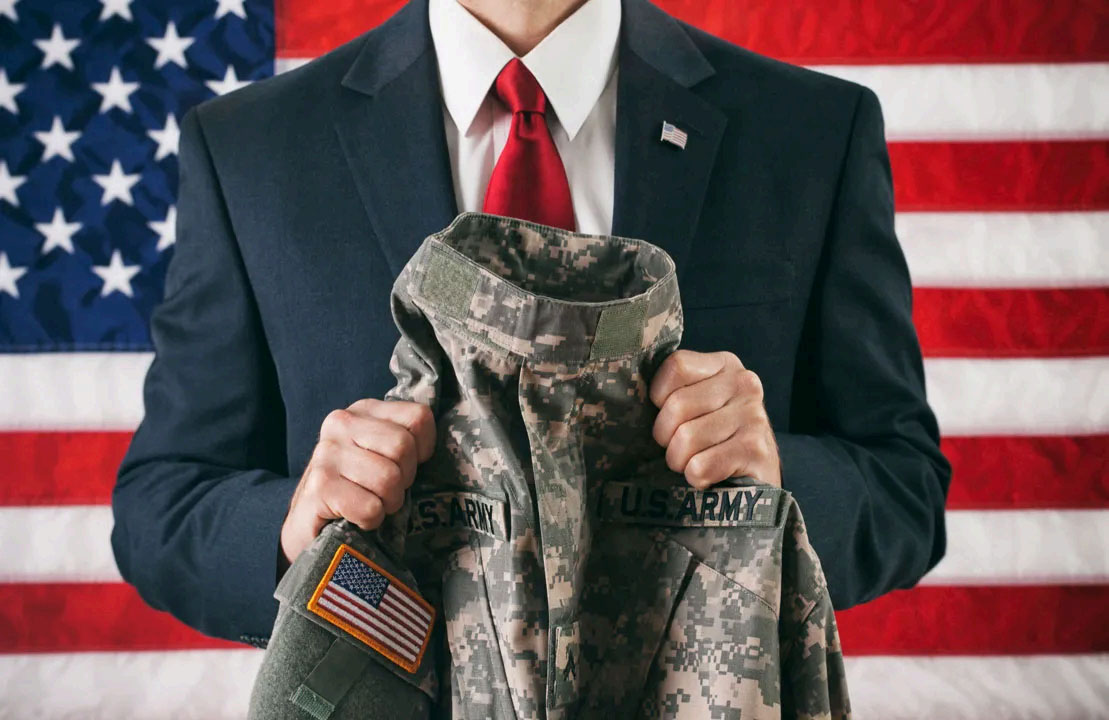 Person in a suit holding a uniform jacket while standing in front of an American Flag.